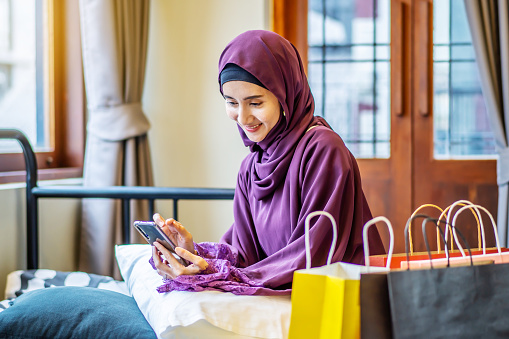 Online shopping concept,Young asian muslim woman sitting on couch in home living room. holding smartphone making online shopping and many shopping bag, browsing online sale website on cellphone