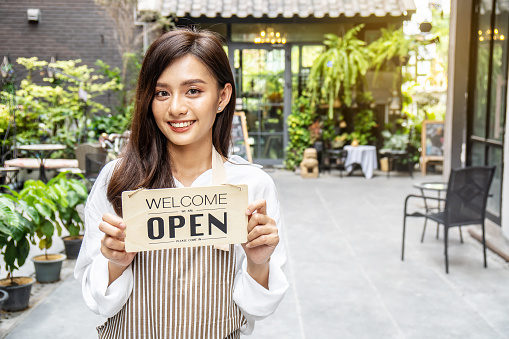Business owner happy beautiful young asian woman in apron looking at camera, we're open sign on front door smiling welcoming clients to new cafe, People and start-up concept.