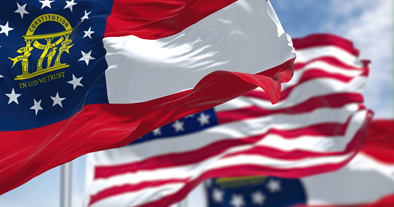 The Georgia state flag waving along with the national flag of the United States of America. In the background there is a clear sky.