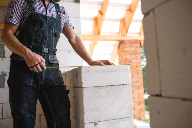 Construction worker at construction site measures the length of window opening and brick wall with tape measure. Cottage are made of porous concrete blocks, work clothes - jumpsuit and baseball cap el trabajador de la construcción en el sitio de construcción mide la longitud de la abertura de la ventana y la pared de ladrillo con cinta métrica. las cabañas están hechas de bloques de hormigón porosos, ropa de trabajo - mono y gorra de béisbol