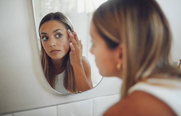 close up of beautiful young caucasian woman looking in the mirror while touching her face and checking for pimples, wrinkles or bags under eyes during her morning beauty routine - look into the mirror imagens e fotografias de stock