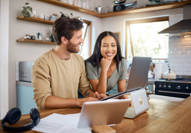 happy young caucasian man working on laptop while his wife stands next to him looking at the screen. man doing freelance work and getting distracted by beautiful wife - rekening stockfoto's en -beelden