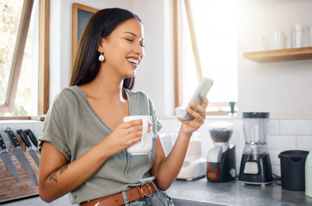 istock Young mixed race hispanic woman using a phone while drinking coffee in the morning at home. Happy woman using social media on her phone standing in the kitchen at home and laughing at funny video 1400838579