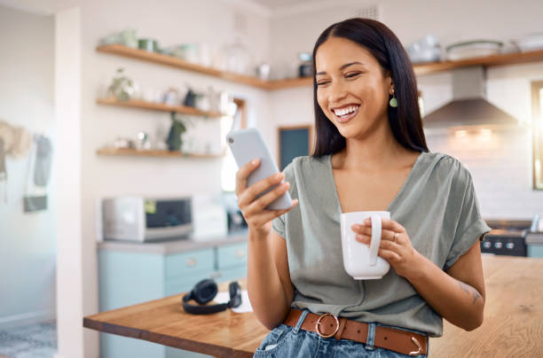 una joven feliz mujer de raza mixta de pie en su cocina en casa y usando el teléfono inteligente para navegar por internet mientras bebe una taza de café. hispano sonriente en las redes sociales y en las redes sociales en un teléfono - usar el teléfono fotografías e imágenes de stock