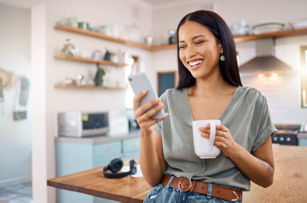 une jeune femme métisse heureuse debout dans sa cuisine à la maison et utilisant un smartphone pour naviguer sur internet tout en buvant une tasse de café. hispanique souriant sur les médias sociaux et réseautage sur un téléphone - portable information device one person social networking text messaging photos et images de collection