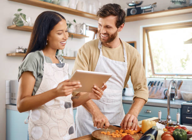 istock Happy young interracial couple using digital tablet while cooking together at home. Young man cutting vegetables while wife reads online recipe. Couple preparing their first meal together 1400838590