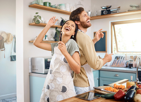 Cheerful young interracial couple being playful singing and dancing in the kitchen while cooking together. Happy young man and woman wearing aprons and having fun at home