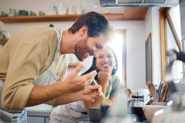 Happy young interracial couple standing by the stove while cooking together in the kitchen at home. Young caucasian man looking and smelling steamy food while it cooks on the stove