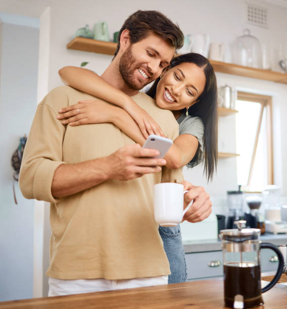 jovem casal jovem feliz sendo amoroso e carinhoso em casa. jovem caucasiano usando seu smartphone e bebe café enquanto sua namorada o abraça por trás enquanto eles olham para a tela digital juntos - dating couple love latin american and hispanic ethnicity - fotografias e filmes do acervo