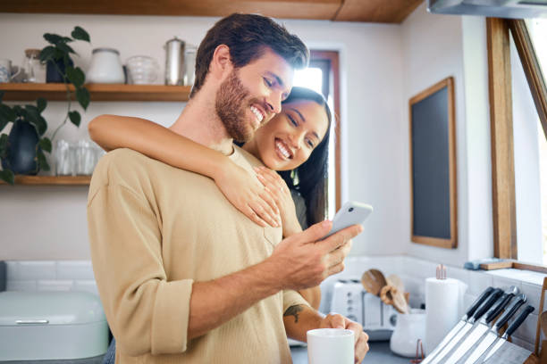 istock Happy young interracial couple being loving and affectionate at home. Young man using his smartphone and holds coffee cup while his girlfriend embraces him from behind. Browsing social media or sending text message 1400838623