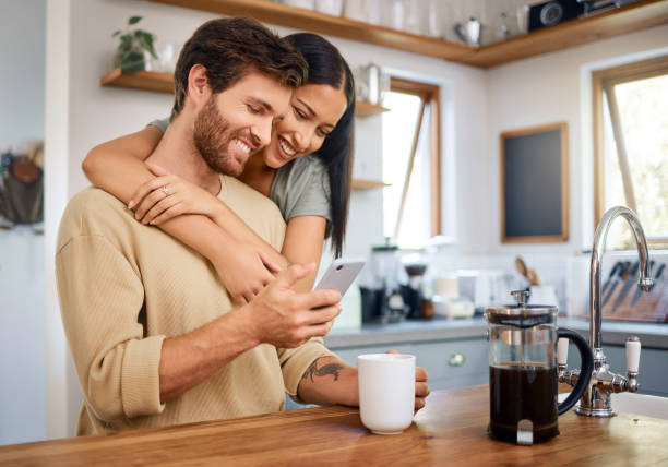 istock Happy young interracial couple being loving and affectionate at home. Young man using his smartphone and holds coffee cup while his girlfriend embraces him from behind. Browsing social media or sending text message 1400838626