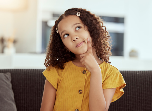 Adorable little mixed race child thinking at home. One small cute hispanic girl sitting alone on a sofa in a living room and coming up with an idea. Bored young kid with curly hair with a solution