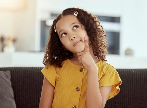 Photo of Adorable little mixed race child thinking at home. One small cute hispanic girl sitting alone on a sofa in a living room and coming up with an idea. Bored young kid with curly hair with a solution