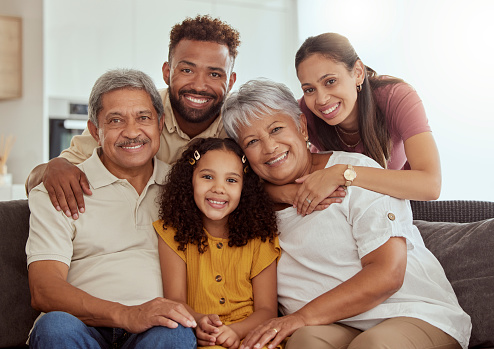 Portrait of mixed race family with child enjoying weekend in living room at home. Adorable smiling hispanic girl bonding with grandparents, mother and father. Happy couples and child sitting together