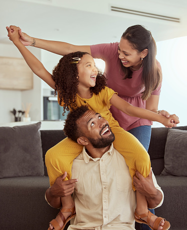 Mixed race parents with daughter playing in living room at home. Adorable smiling hispanic girl on father’s shoulders and bonding with mother while pretending to fly. Happy couple and child together