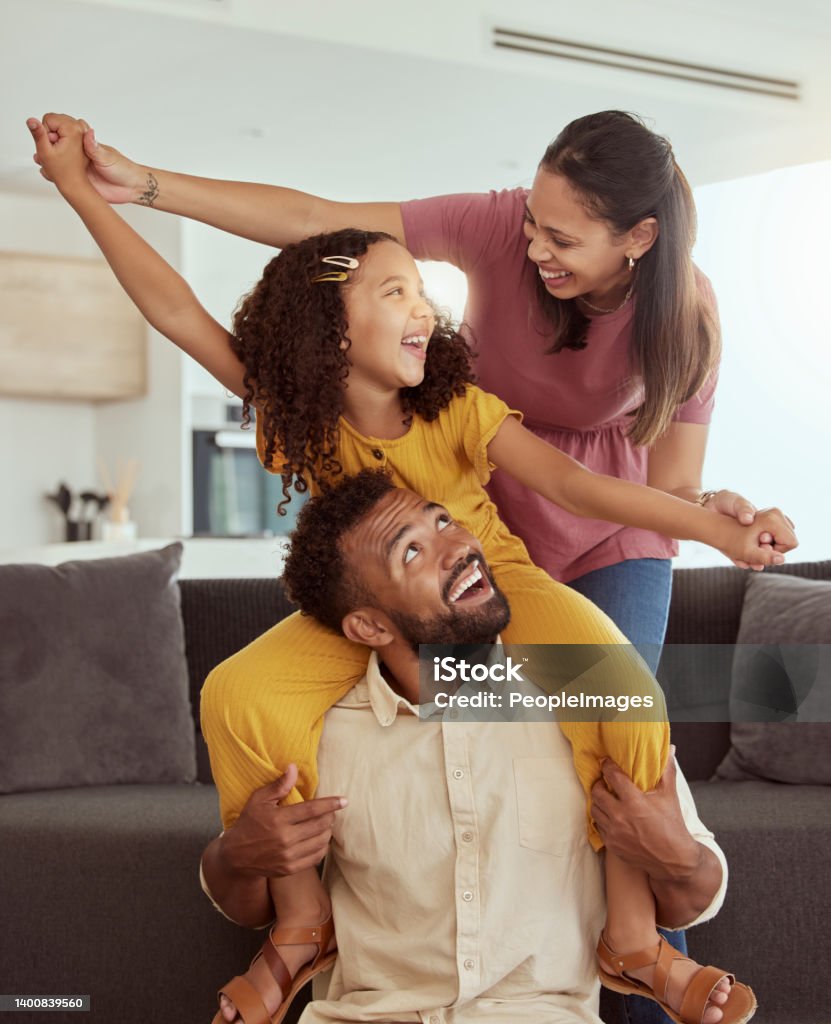 Mixed race parents with daughter playing in living room at home. Adorable smiling hispanic girl on father’s shoulders and bonding with mother while pretending to fly. Happy couple and child together Family Stock Photo
