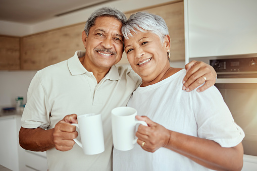 Portrait of mixed race senior couple enjoying morning coffee at home. Smiling elderly husband and wife standing together and drinking tea in kitchen. Happy retired man and woman hugging and bonding