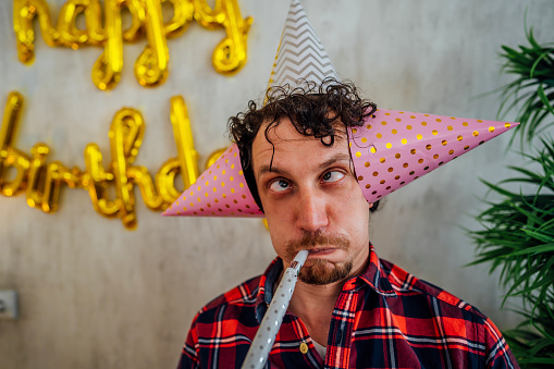 Young funny man blowing party horn during birthday celebration and making silly faces