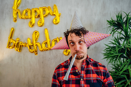 Young funny man blowing party horn during birthday celebration and making silly faces