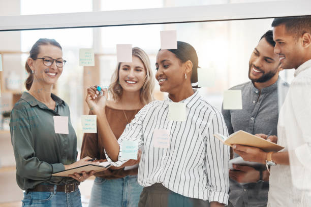 istock Confident young mixed race businesswoman writing notes and planning a project on a glass wall in an office. Group of smiling businesspeople brainstorming ideas and strategies together in a creative startup agency 1400842076