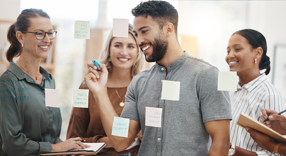 Confident young mixed race businessman writing notes and planning a project on a glass wall in an office. Group of smiling businesspeople brainstorming ideas and strategies together in a creative startup agency