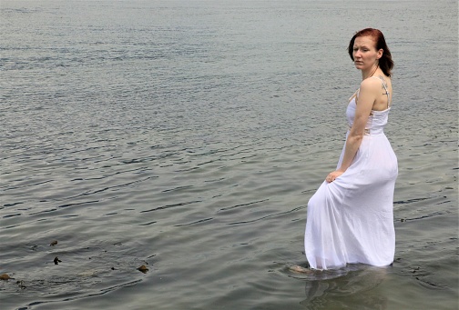 A Caucasian model wading in the Pacific Ocean. She is wearing a long, sleeveless, white dress.