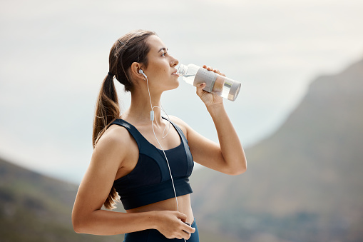 One fit young mixed race woman taking a rest break to drink water from bottle while exercising outdoors. Female athlete wearing earphones quenching thirst and cooling down after running and training workout