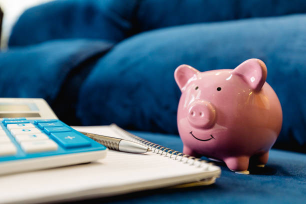 青いソファに掛けられた貯金箱には、電卓、開いたノート、紙幣用のペンが置いてあります。ホームファイナンスの概念、予算と経費。 - currency pen wealth paper currency ストックフォトと画像