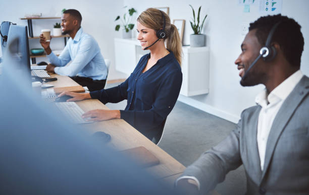 istock Happy young caucasian call centre telemarketing agent talking on a headset while working on computer alongside colleagues in an office. Confident friendly female consultant operating helpdesk for customer service and sales support 1400849587