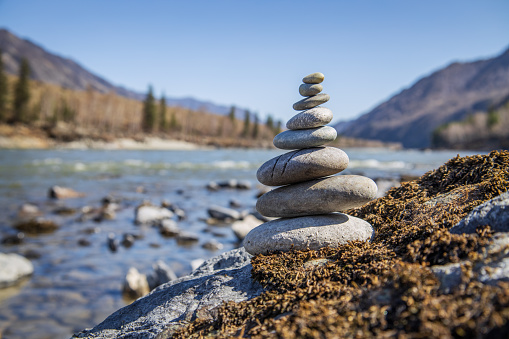 Pyramid of stones for meditation and making wishes on the bank of a mountain river against the backdrop of mountain peaks and forest. Horizontal landscape in cold shades.
