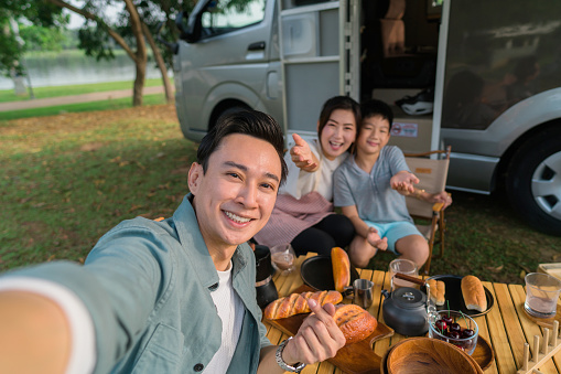 happy family traveling in campervan and picnic outdoor, spending time together while father taking selfie using smart phone, concept of motor home.