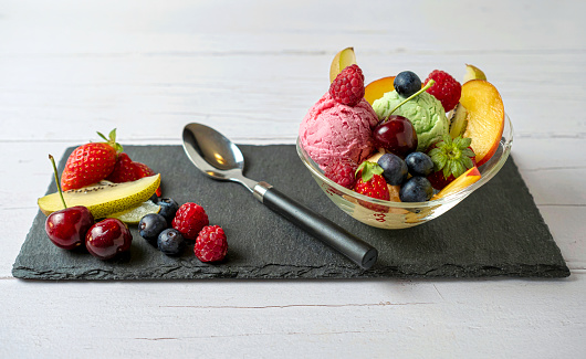 Fruit salad with scoop of vanilla ice cream, strawberry, pistachio in a bowl on a piece of slate, isolated on white background.