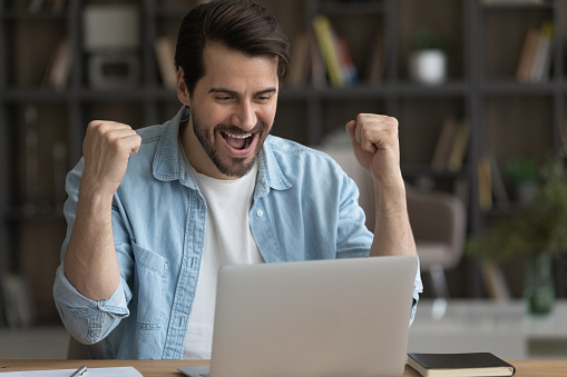 Triumphant. Excited young businessman working from home office raise fists shout yes yeah by laptop receiving credit on affordable terms. Joyful millennial male getting great contract business victory