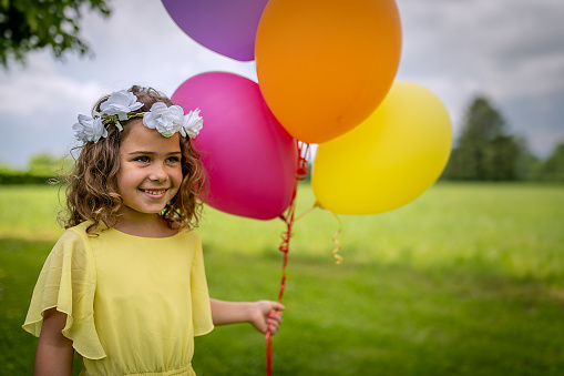 A smiling girl in spring nature in a yellow dress, with a wreath of flowers on her head and with colored balloons filled with helium.