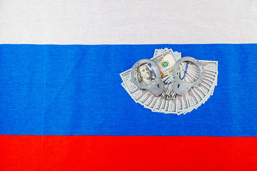 silver metal police handcuffs over paper dollar banknotes of United States of America over national flag of Russian Federation, concept of foreign currency prohibition in high angle view