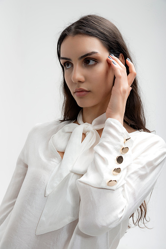 The young and beautiful photomodel wearing a thin fabric, summer white shirt and white pants poses artistically in the studio with a white background. Photo shoots for the clothing industry, which is a textile product for e-commerce. Image work. Cover images. A brunette, wheat-skinned young girl with long dark hair.