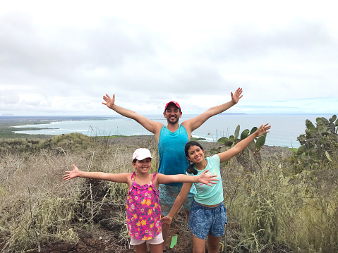 Vacation Portrait of a latin family: A young latin father, and two daughters looking at the camera while enjoying their vacation time in Galapagos Island, Ecuador in a cloudy day, over a hill with Isabela town and the rest of the island on back.

Travel Concept.
Idyllic touristic Travel Destination Concept.