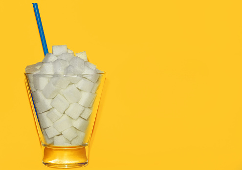Glass cup with sugar on a yellow background, the concept of a harmful drink with a lot of sugar, energy in a glass, blue straw, white sugar cubes