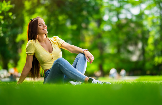 Beautiful young woman relaxing with eyes closed  while sitting on grass in the park. Vibrant colors on beautiful spring day.