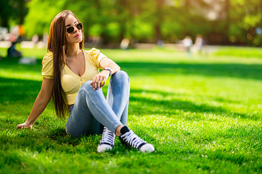 Beautiful young woman sitting on grass and relaxing in the park. Vibrant colors on beautiful spring day.