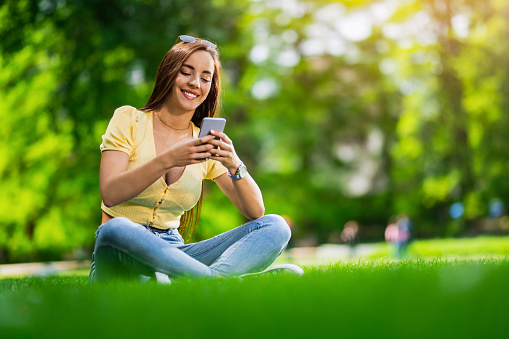 Beautiful young woman text messaging on her smart phone in the park. Vibrant colors on beautiful spring day.