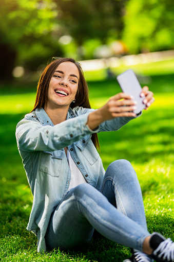 Beautiful young woman taking a selfie with smart phone while sitting on grass in the park. Vibrant colors on beautiful spring day.
