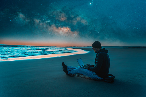 person sitting with laptop on the beach outdoors working under the starry night and milky way