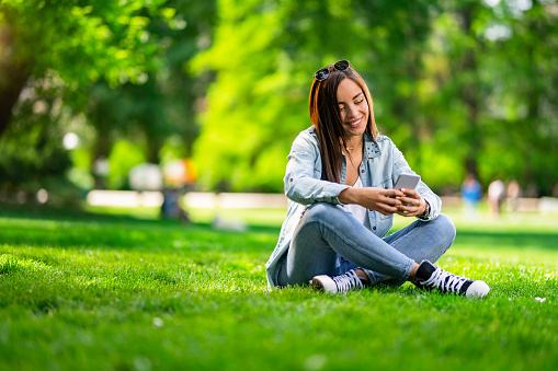 Beautiful young woman text messaging on her smart phone while sitting on grass in the park. Vibrant colors on beautiful spring day.