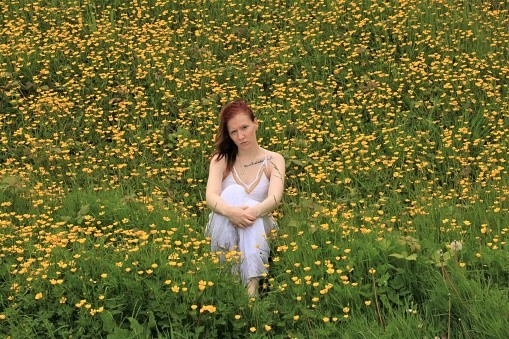 A tattooed Caucasian woman sitting in a patch of wild Buttercups. She is wearing a white long sleeveless dress.