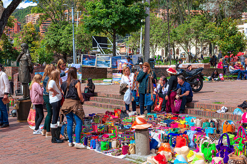 Bogotá, Colombia - May 21, 2017: The weekly Sunday event of the Mercado de Las Pulgas or Flea Market in the historic Usaquen District of the capital city, is in full swing. Some tourists look at brightly colored Mochila bags for sale. To the extreme left of the image, there is a mime artist standing still as a statue. For a few pesos he will move to thank you. The focus of the Flea Market is on enjoyment: there is music, entertainment, shopping and a variety of food including various snacks and deserts, available. The altitude at street level is about 8,6600 feet above mean sea level. Photo shot in the afternoon sunlight; horizontal format.