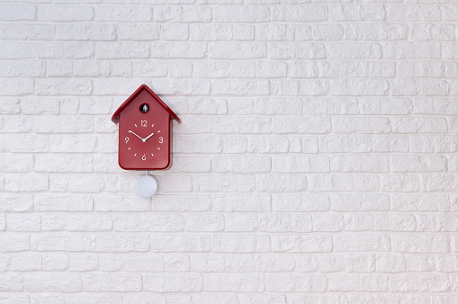 White cuckoo bird clock with red round pendulum and dial on a white brick wall with copy space