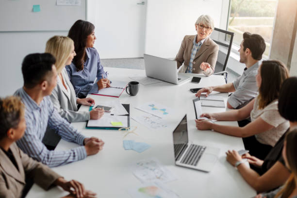 Senior businesswoman with group of multiracial work colleagues discussing ideas and business together sitting at table in modern creative office interior high angle view daytime Senior businesswoman with group of multiracial work colleagues discussing ideas and business together sitting at table in modern creative office interior high angle view daytime corporate hierarchy stock pictures, royalty-free photos & images