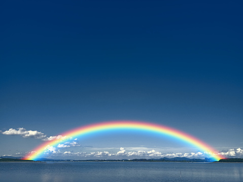 Rainbow on dark blue sky and white cloud on lake and line of tree and tiny mountain background