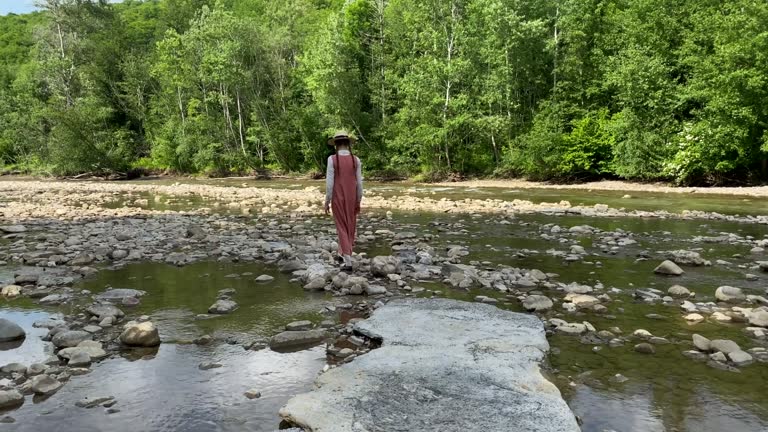 Retro styled young woman in straw boater hat and long dress walking on river rocks near forest. Romantic girl. Natural scenic landscape. Summer vacation. Vintage aesthetic lifestyle. 4K video